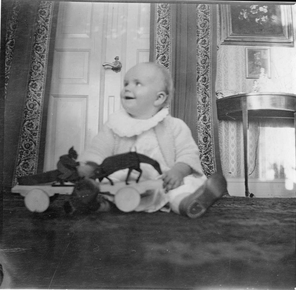 Foto av ukjent baby sittende på gulv med leker. Antagelig et av Aall barna Cecilie Aall gift Mathiesen, Niels Frederik Aall eller Cato Aall