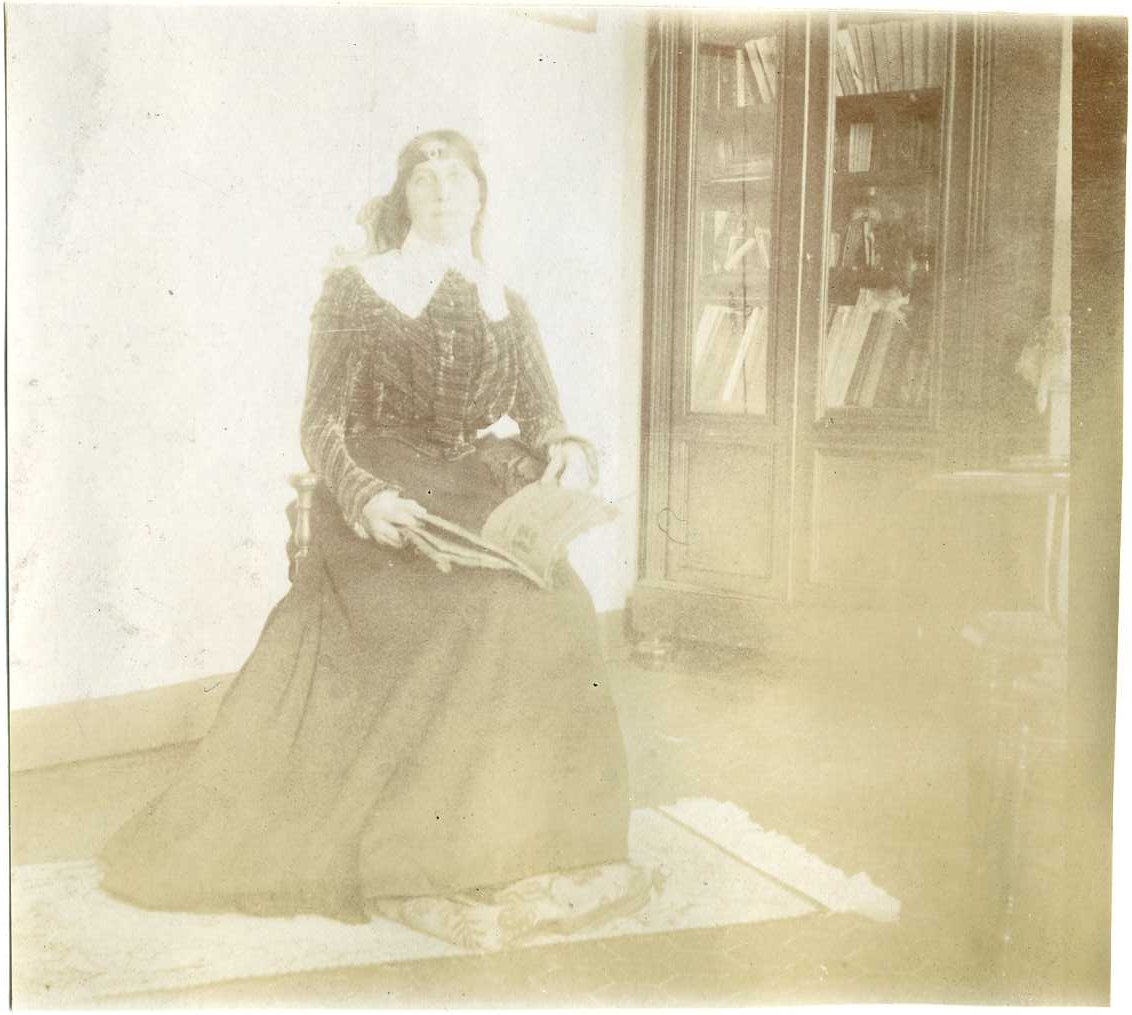 Porträtt av en kvinna som sitter med en uppslagen bok i knät vid ett bokskåp. Hon bär ett pannband. "Mademoiselle Maxim"?
