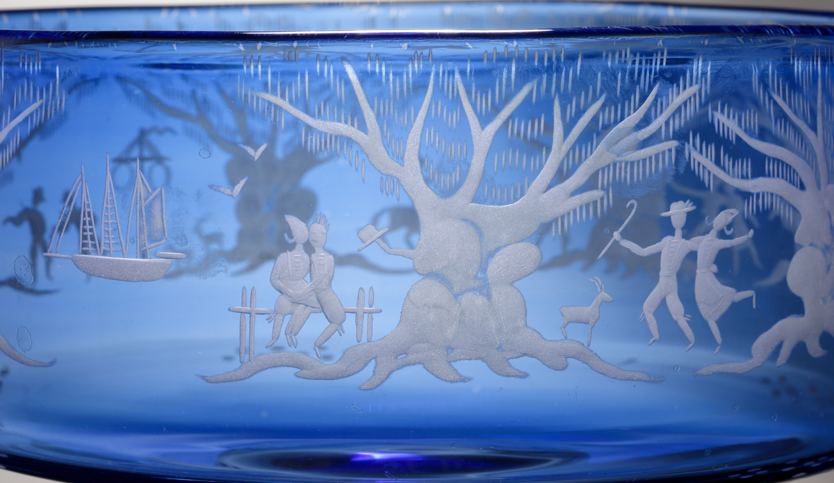Formgivare: Gunnar Cyrén. Opalblå skål med cylindrisk form och rund ofärgad klackfot.
Runt livet graverat naturmotiv med ekar, en midsommarstång och dansande och spelande människor.