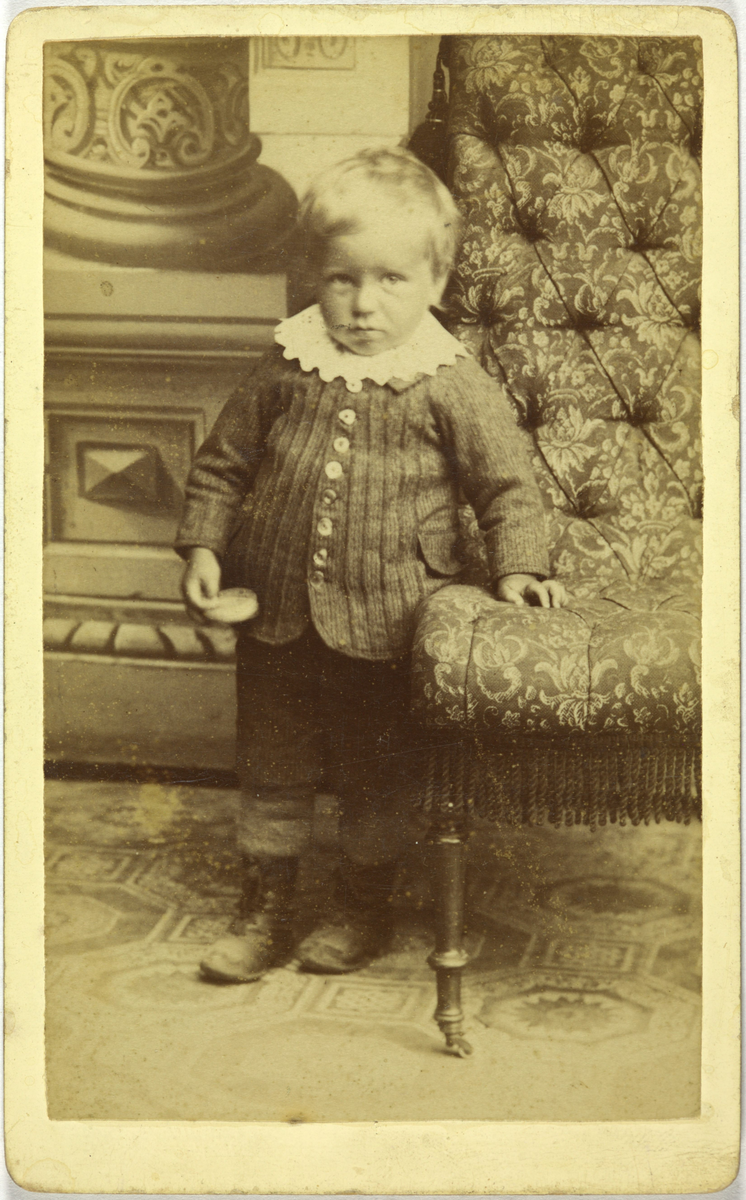 Atelierfoto av ein liten gut, han står ved en lenestol.