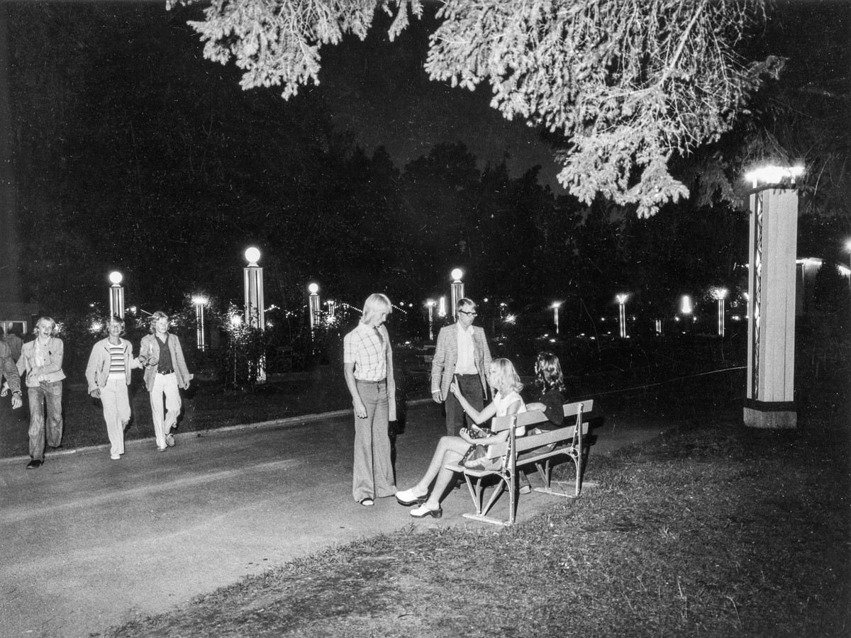 Utomhus- och kvällsbild från parken i Folkets Park i Linköping, 1970-tal. I förgrunden en parkbänk där två kvinnor sitter, framför dom står två män. I bakgrunden till vänster kommer några personer gående.