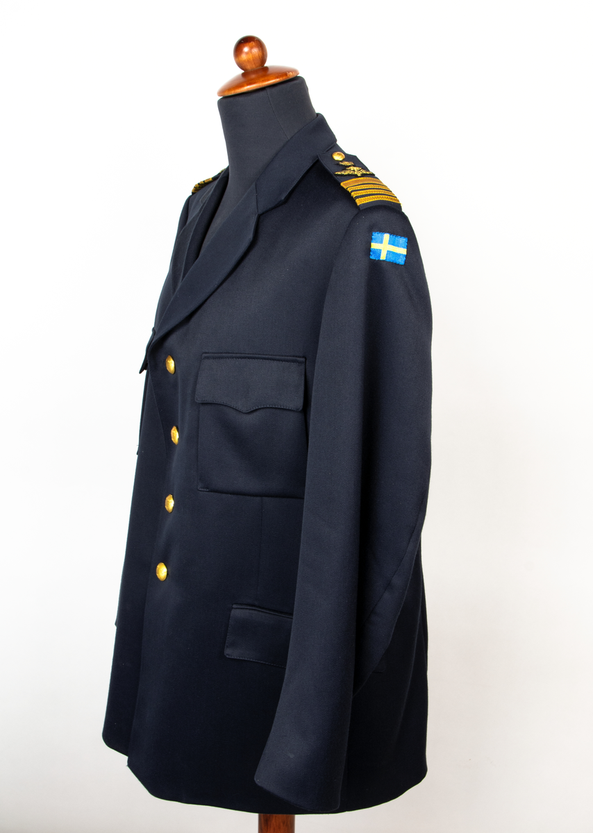 Vapenrock m/87. Rock av blått tyg, knäpps med fyra uniformsknappar, försedd med fasta axelklaffar och ficklocken är formade.
Axelklaffarna är försedd med påsydd gradbeteckning, överste 1 graden. På vänster överarm är en nationsbeteckning i form av en svensk flagga fastsydd och på höger bröstficka sitter försvarsstabsmärke m/49.