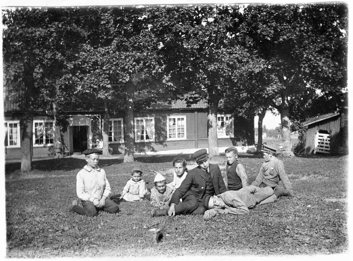 Personer på plenen foran hovedbygningen på Evenrud, Kapp i Østre Toten, 1904-1905. Personene er: Thorvald Raabe, Aagot Sveinson (f.Raabe), Sverre Raabe (med papirhatt), Pauline Hveem (f.Raabe), Petter Bekkevold (Appelsvollsveen), Karl Moen (kalte seg senere Sandvoll), Frantz Raabe (liggende foran).