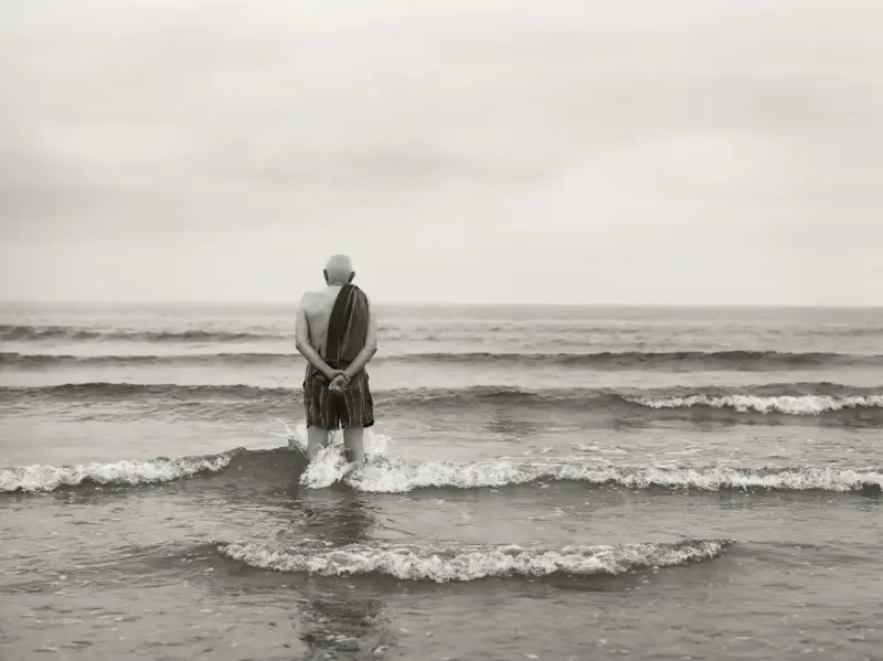 En mann står ute i vannet, han har på shorts og har et håndkle over skuldra. Utsikten er kun hav.