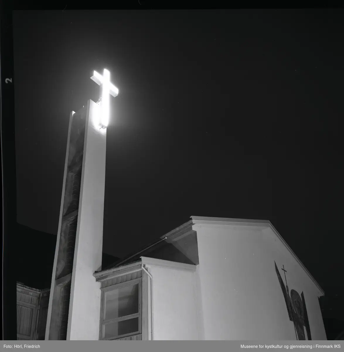 Det lyssatte korset ved klokketårnet til den katolske kirka St. Mikael i Hammerfest skinner i nattemørke. Deler av mosaikken på fasaden er synlig nede til høyre i bilde. Fotoet er tatt oppover fra gateplan i Strandgata.