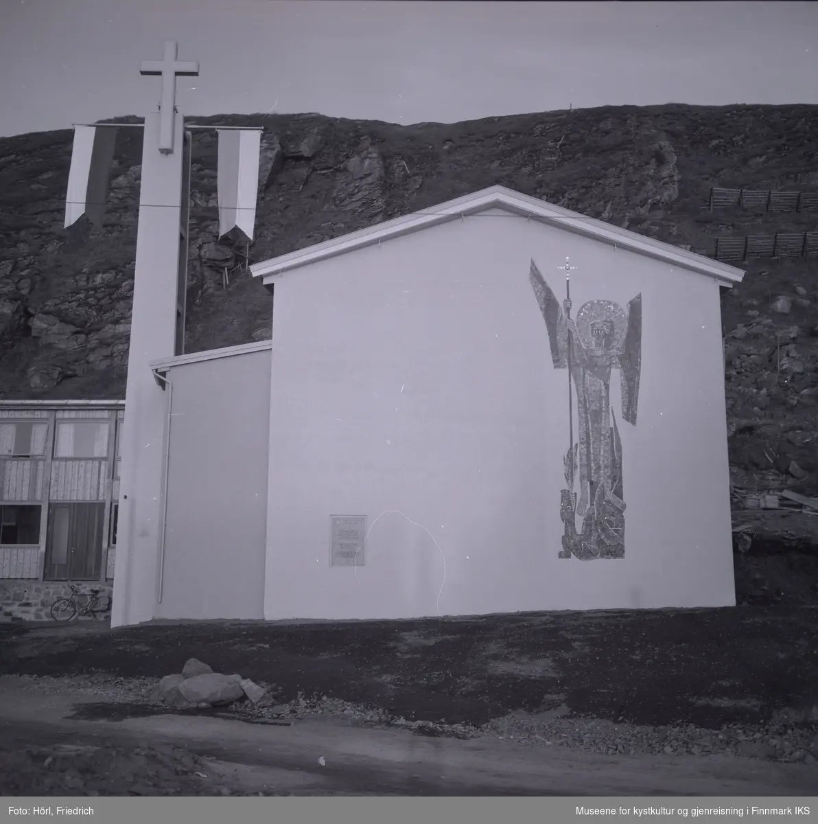 Ved klokketårnet til den katolske kirka St. Mikael i Hammerfest er det montert to stenger med vimpler. Det antas at dette ble gjort i anledning vigslingen den 3. august 1958. 
Kirkens vegg mot gata er dekorert med en stor mosaikk av St. Mikael, skapt av den tyske billedkunstneren Karl Manninger. Ved kirkens sidefløy til venstre er en sykkel lent mot veggen. Bak kirkebygget ser man Salenfjellet.