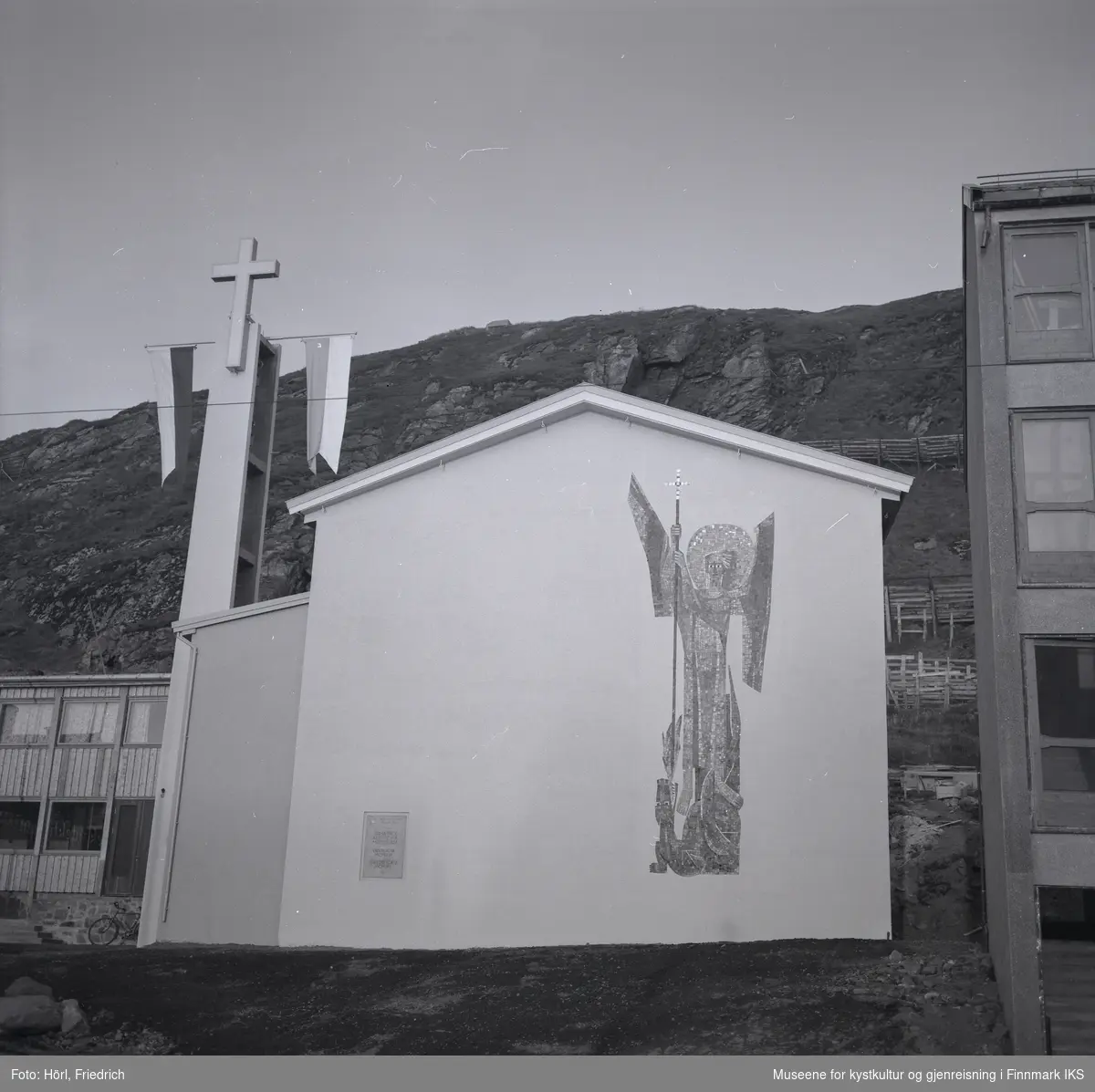 Ved klokketårnet til den katolske kirka St. Mikael i Hammerfest er det montert to stenger med vimpler. Det antas at dette ble gjort i anledning vigslingen den 3. august 1958. 
Kirkens vegg mot gata er dekorert med en stor mosaikk av St. Mikael, skapt av den tyske billedkunstneren Karl Manninger. Ved kirkens sidefløy til venstre er en sykkel lent mot veggen. Bak kirkebygget ser man Salenfjellet. På høyre bildekant ser man deler av nabobygget.