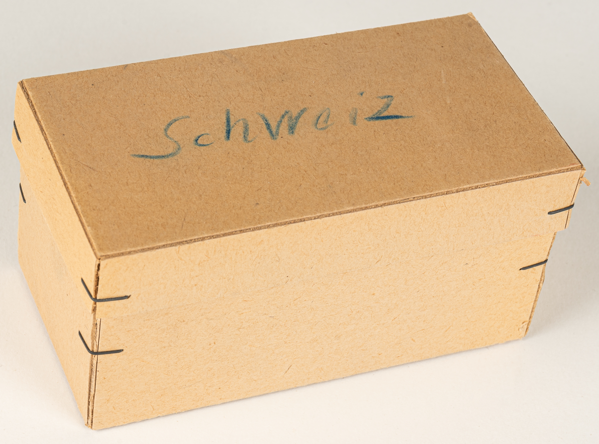 Ask i grå papp, innehållande fyra tablettaskar i plåt, gröna Läkerolaskar. På pappasken handskriven text "Schweiz". Den ena tablettasken har öppning på baksidan för montering.
Läkerolaskar med text på tyska.