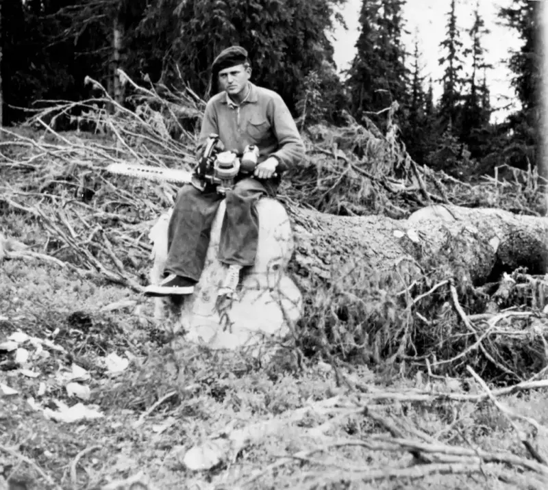 Svart-hvitt foto av mann som sitter på en stor tømmerstokk som akkurat er felt, han har motorsag i fanget.