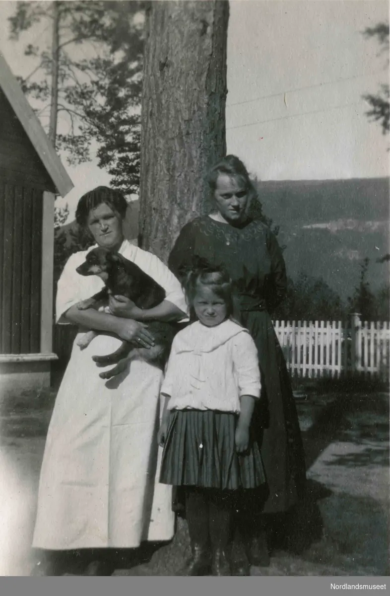 To kvinner og et barn står foran et stort tre. Den ene kvinnen holder en hund.