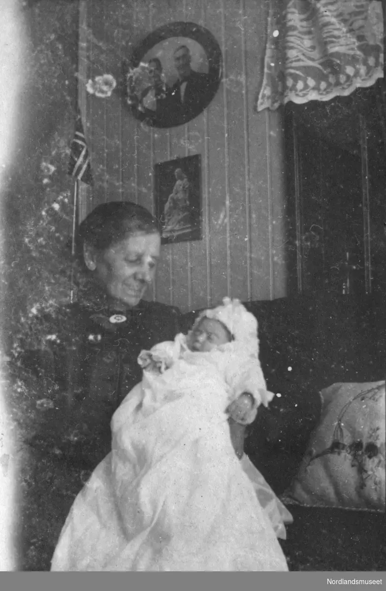 En eldre kvinne holder et spedbarn i dåpskjole. Interiør.