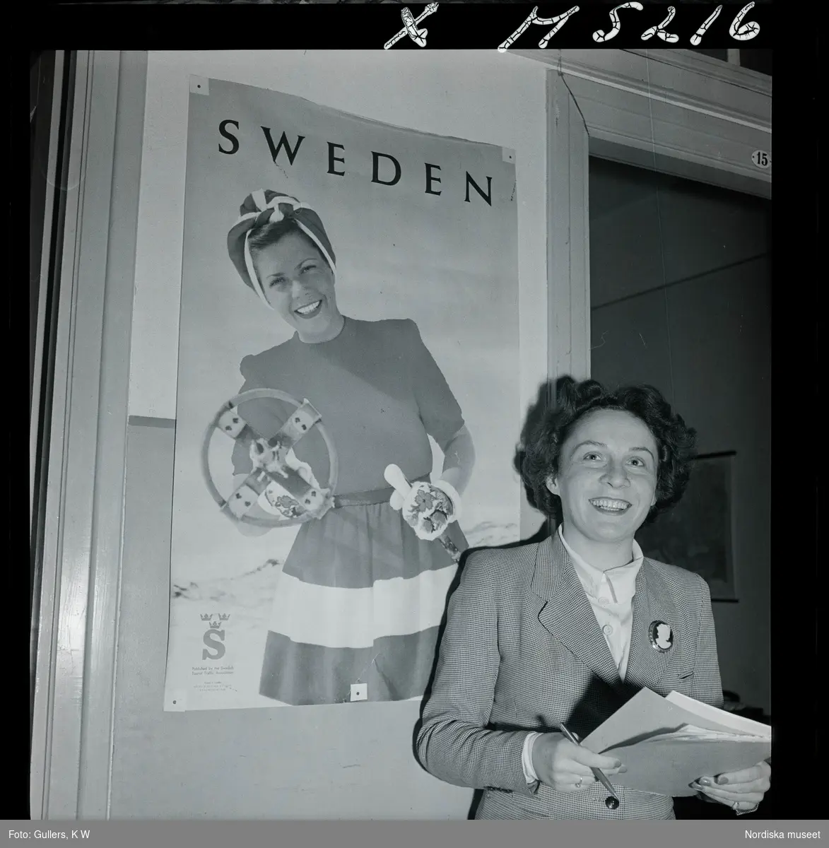 1717/K Istanbul allmänt. En leende kvinna med papper i händerna står framför en affisch som gör reklam för Sweden.