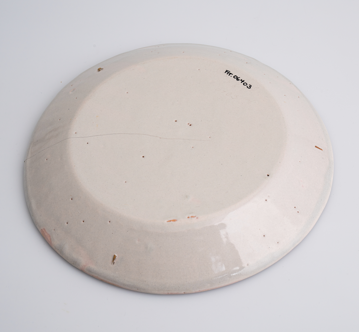 En flat tallerken av keramikk/porselen. Den har bly- og tinnglasur og er hvit på farge. Tallerkenen er rund med en relativt høy og skrånende kant. Den har ikke dekor utover glasuren. Tallerkenen er en av 11 identiske tallerkener som trolig er fra tidlig på 1700-tallet.

