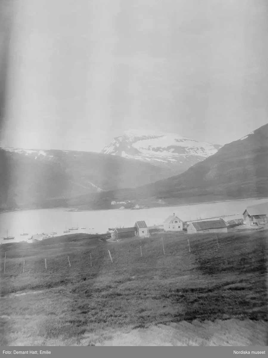 Vy över bebyggelse, åkrar, vatten och fjäll i Norge. Bygden som idag är en förort till Tromsø heter Hungeren. Bilden ingår i en serie fotografier tagna av Emilie Demant Hatt i Sapmi mellan åren 1907 och 1916.