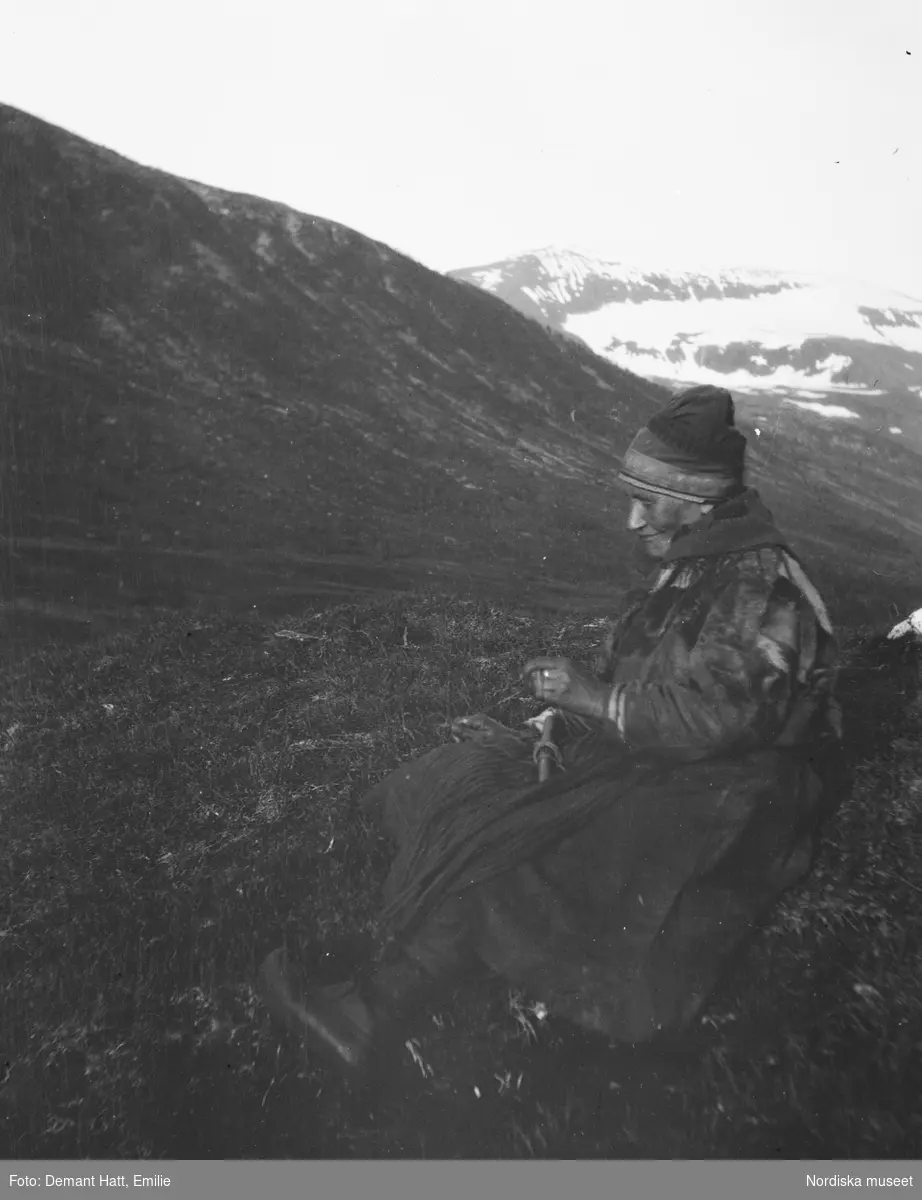 Kvinna, Girste, enligt bildtext gift med Andaras, sitter i landskap med fjäll i bakgrunden och handarbetar. Bilden ingår i en serie fotografier tagna av Emilie Demant Hatt i Sapmi mellan åren 1907 och 1916.