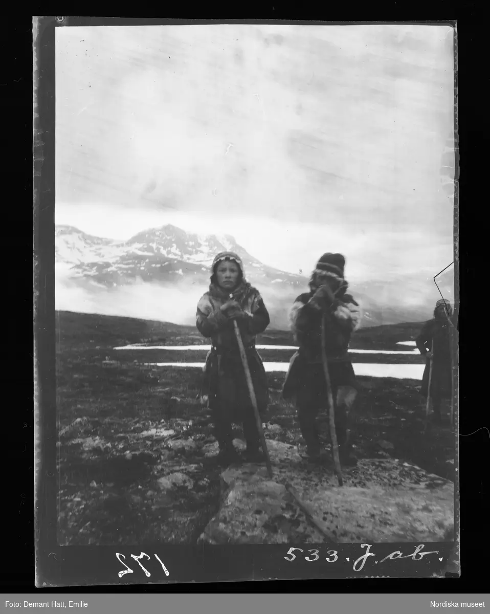 Tre barn/ungdomar med vandringsstavar ute i naturen. I bakgrunden fjäll. Vårflyttningen från Närvä. Bilden ingår i en serie fotografier tagna av Emilie Demant Hatt i Sapmi mellan åren 1907 och 1916.