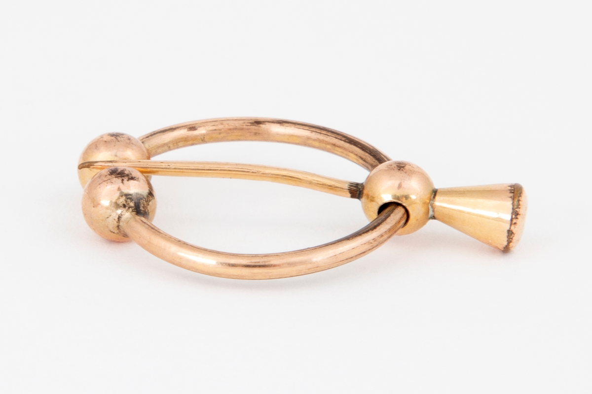 Brosje i gulldublé formet som en åpen ring med to kuler i hver ende. En bevegelig nål med kjegleformet hode er festet til ringen med en tilsvarende kule.