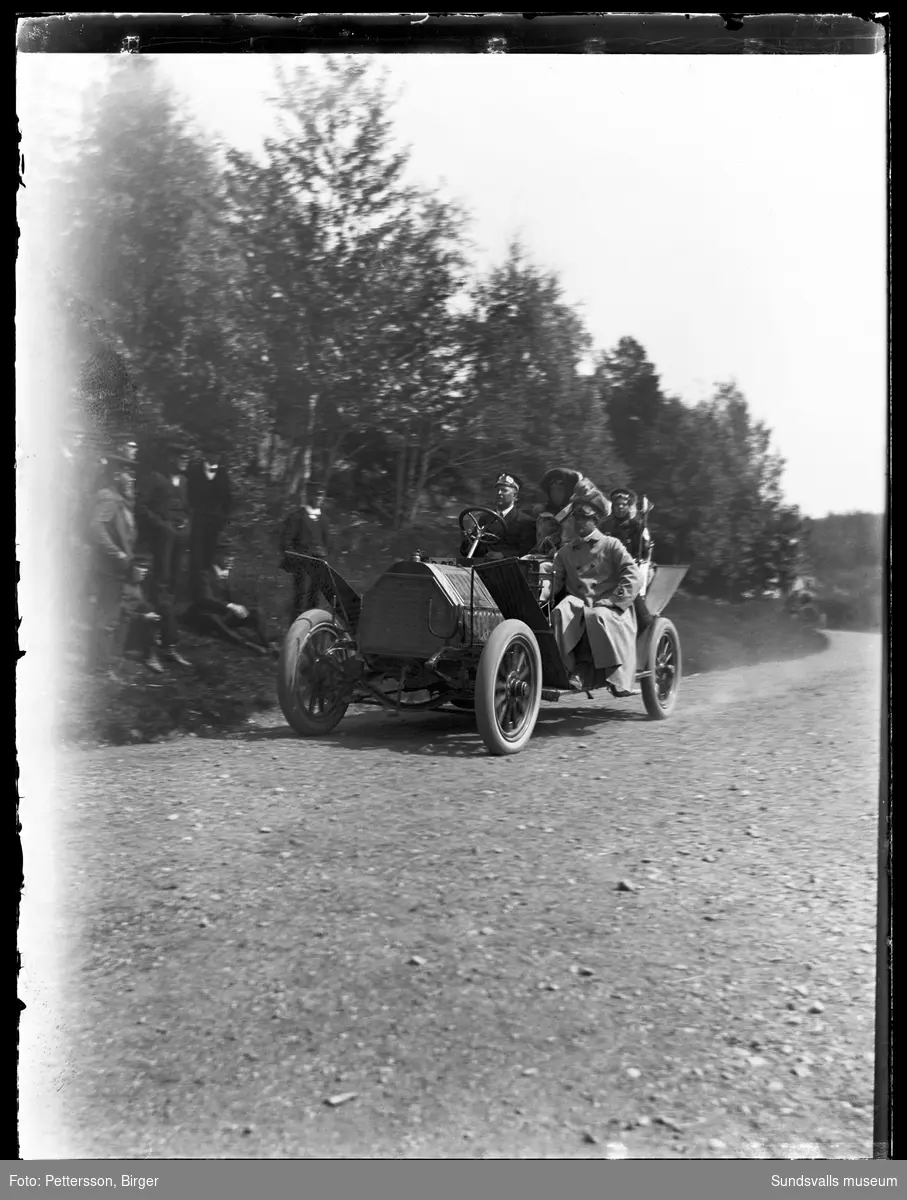En ålderdomlig öppen bil utan vindruta på en landsväg, troligen i Njurundatrakten. I bilen sitter fyra personer och vid vägkanten står en grupp män och beundrar fordonet. Bilden är tagen vid en biltävling som Svenska Motorklubben (SMK) anordnade sommaren 1909 på sträckan Haparanda-Stockholm. Bilen är en 40 hästkrafters Horch från 1907 som ägdes av firman Bil-Bolaget.