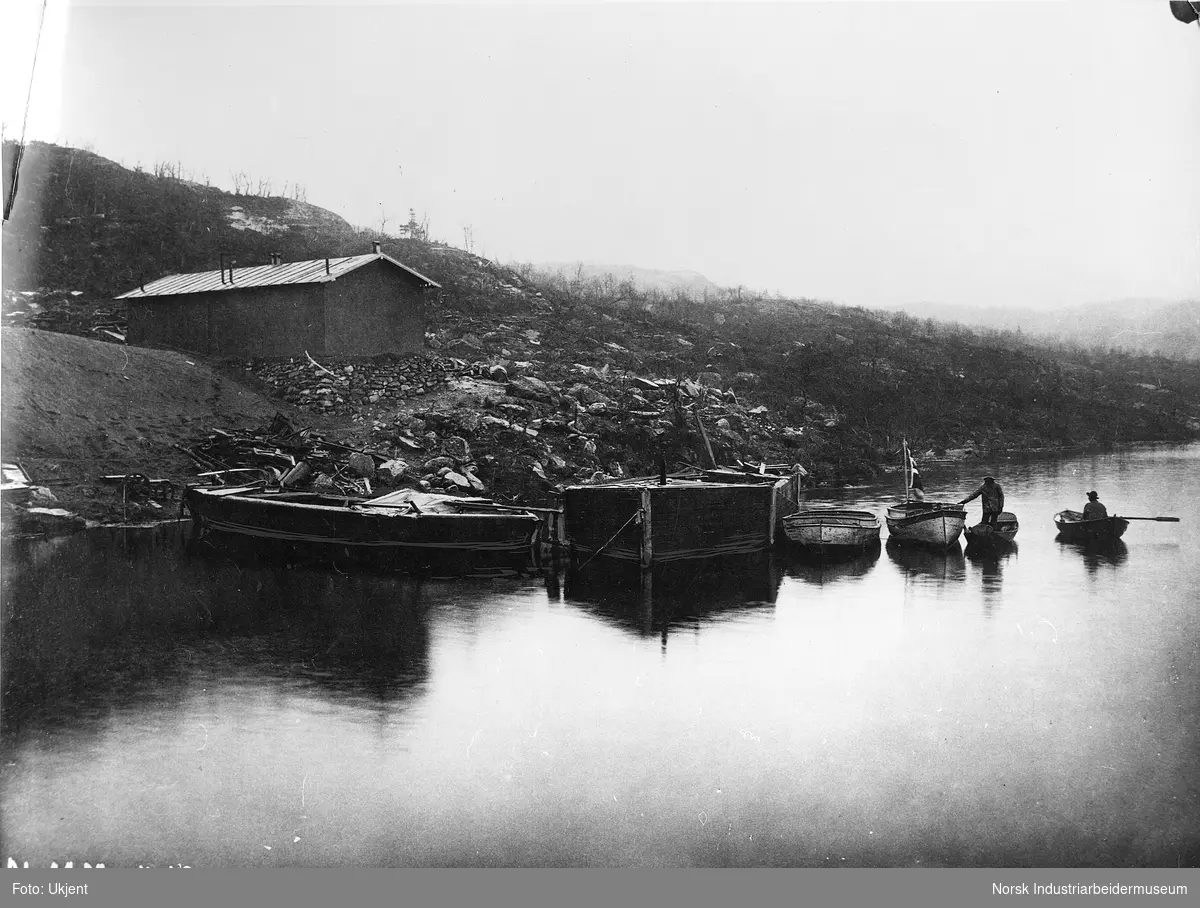 Småbåter og lektere på Møsvann brukt i forbindelse med bygging av Møsvannsdammen. To robåter med menn i, en båt med norsk flagg. Brakke i bakgrunnen.
