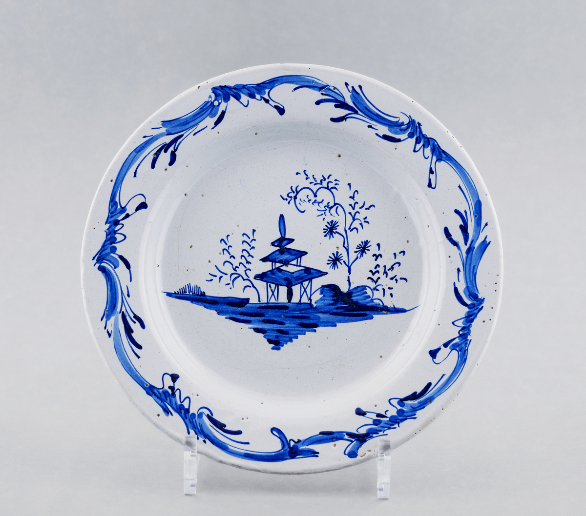 Fat / stor tallerken, flat med hvit glasur, dekorert i dyp blått med pagode og trær i bunnen, rokokkobord langs kanten.