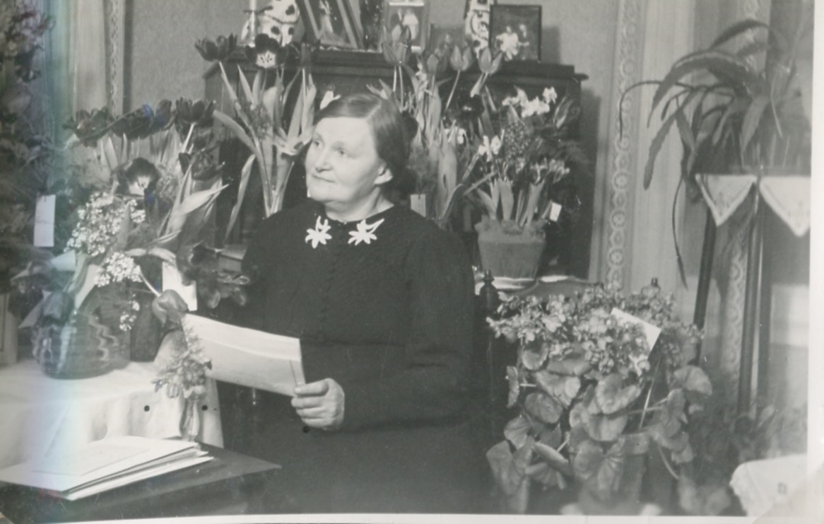 Svea* Gudviva Fredrika Johansson(1892 - 1976) sitter omgiven av blommor. Hon håller ett papper i handen, Heljered Västergård "Brors", okänt årtal. Svea var söndagsskolärare i Missionsförsamlingen.
