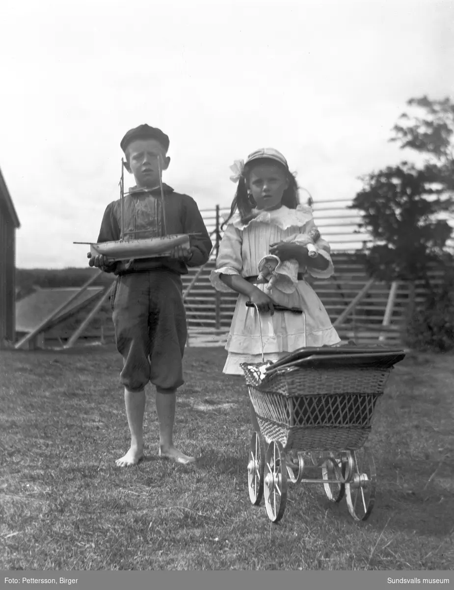 En pojke och en flicka med sina leksaker, pojken har en segelbåt och flickan har docka och dockvagn. Bakom syns en storhässja samt lador.