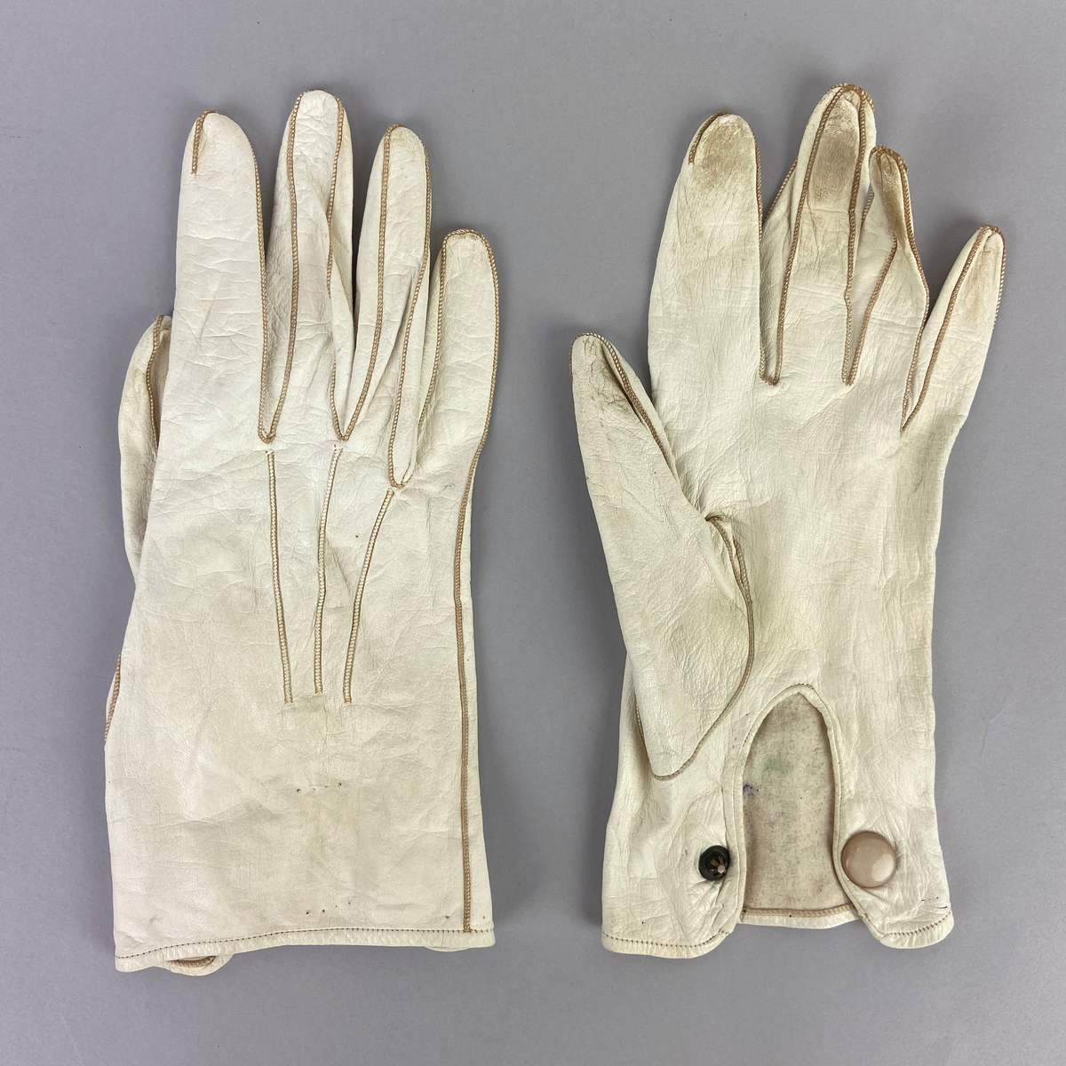 Handskar, tunna vita i glacéskinn. På översidan av handryggen är tre sömmar och på undersidan vid handleden knäpps handskarna med en vit knapp. Avsedda för trupparad och högtidsdräkt, sommartid