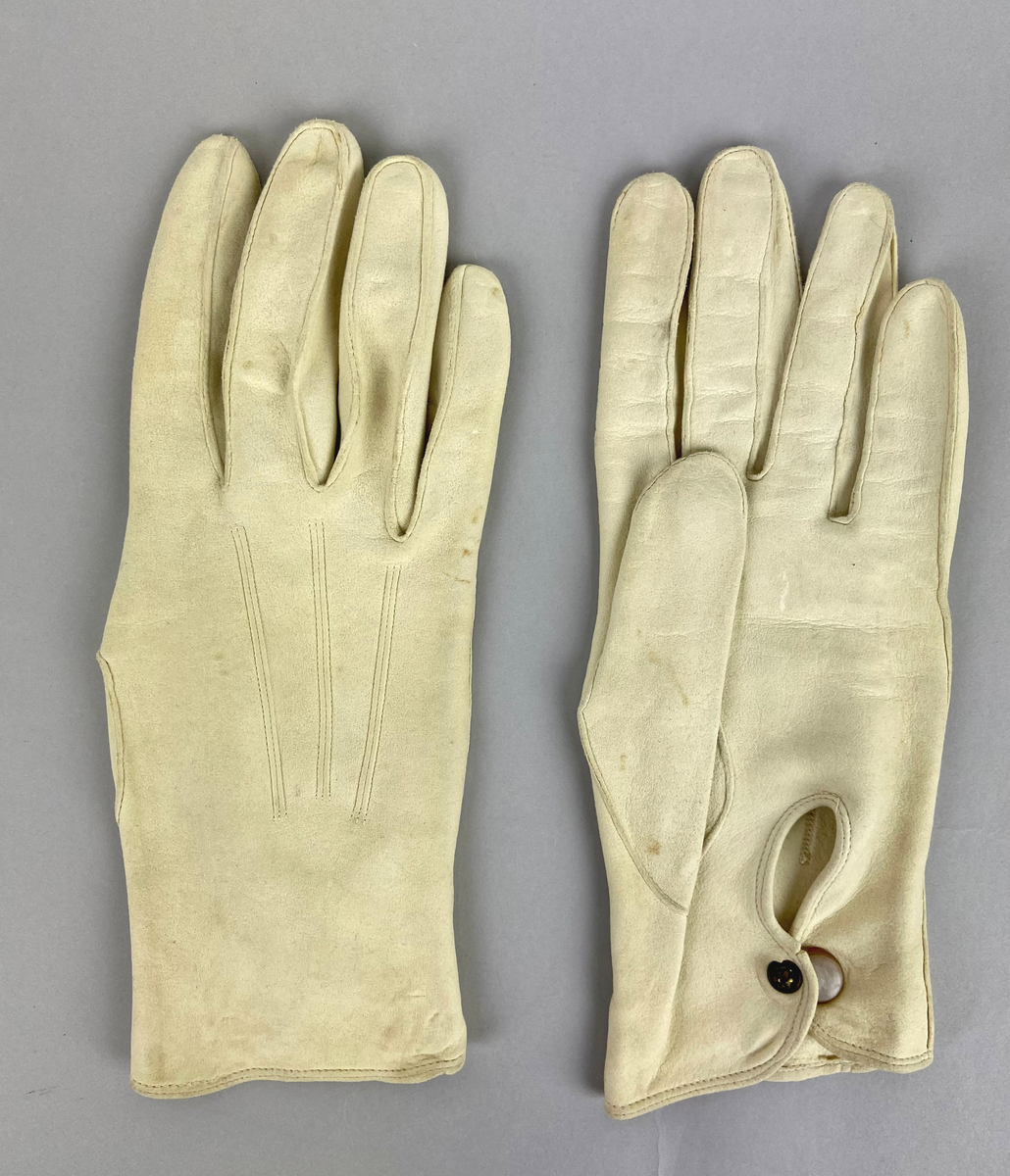 Handskar, tunna vita i sämskskinn. På översidan av handryggen är tre sömmar och på undersidan vid handleden knäpps handskarna med en vit knapp. Avsedda för trupparad och högtidsdräkt, sommartid