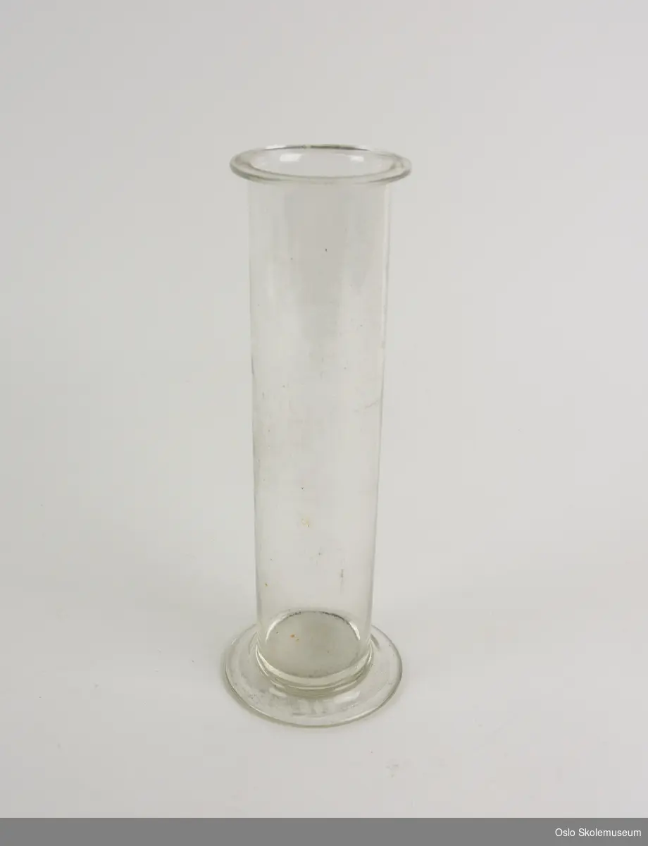 Sylinder i glass med utbrettet base nederst. Øverst er glasset formet i en liten tut. På den ene siden er det en måleskala i hvitt fra 10 til 250.