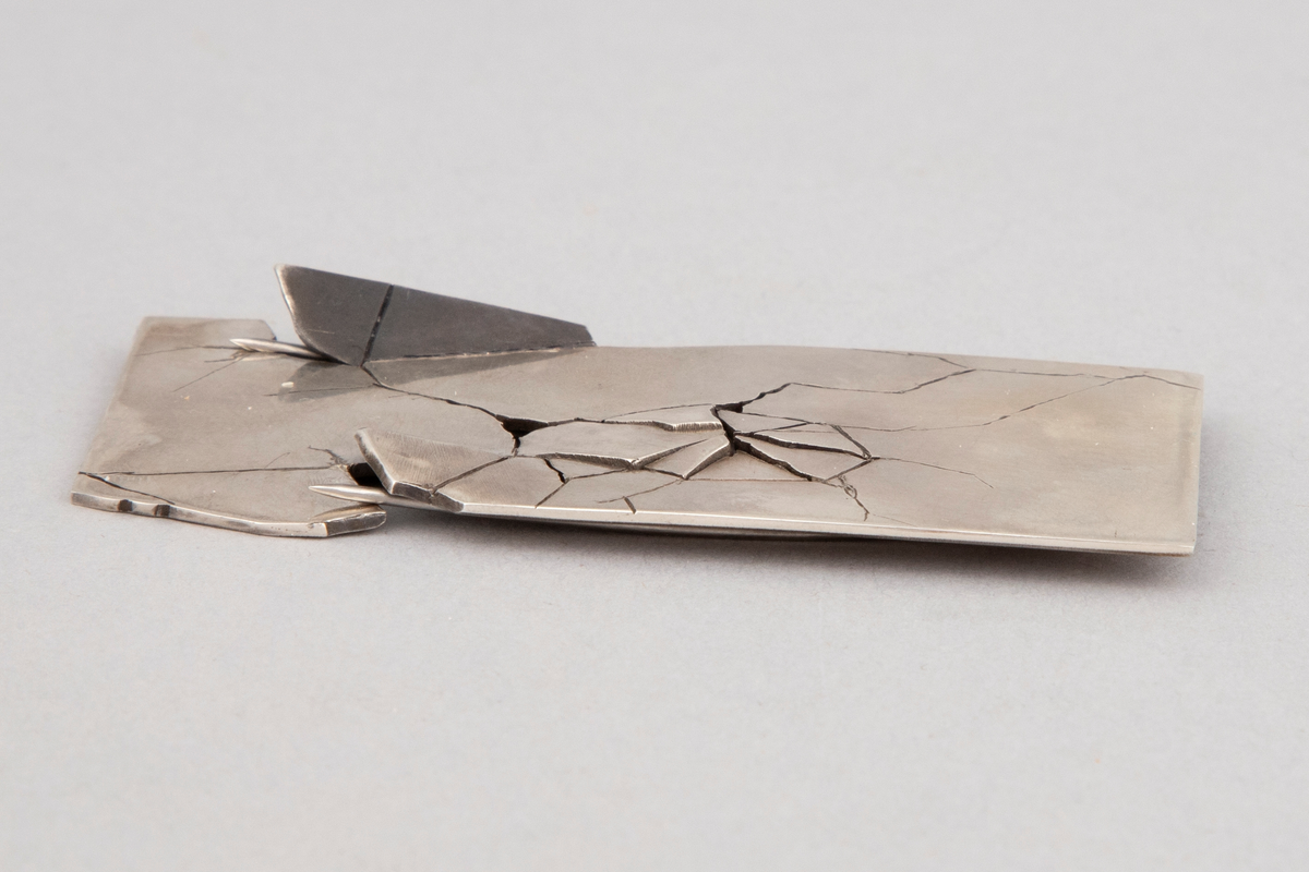 Rektangulær brosje i sterling sølv med sprekker og brudd på framsiden. Dobbel nål på baksiden.