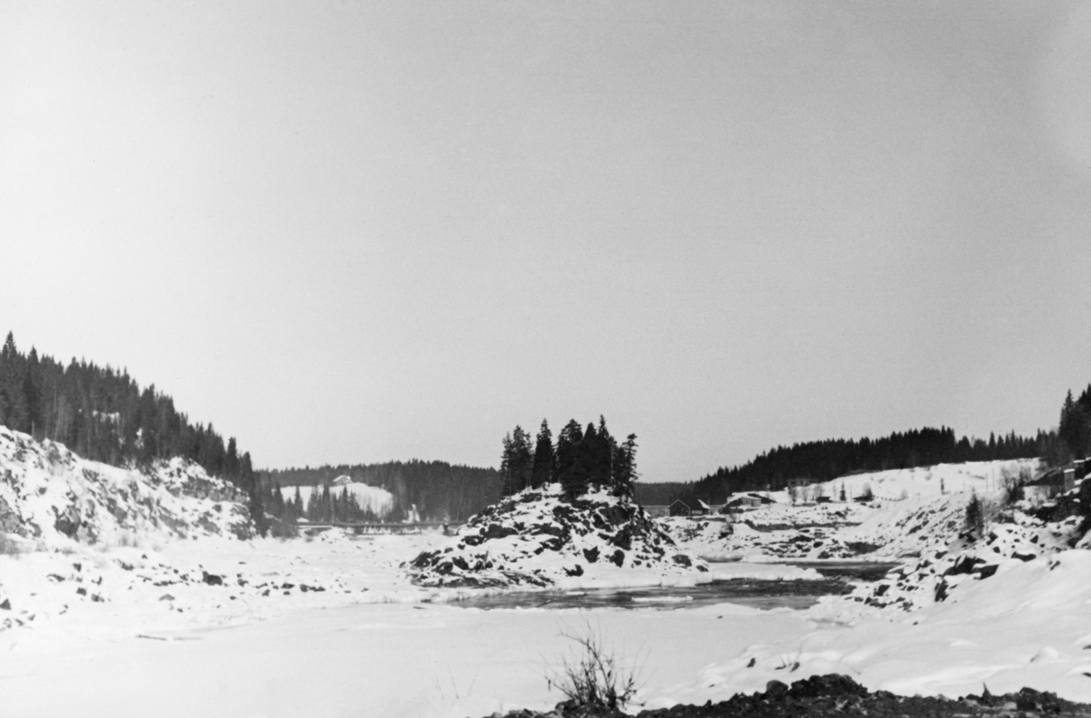 Elvelandskap ved Kykkelsrud i Nedre Glomma, på grensa mellom Askim og Spydeberg kommuner, fotografert vinteren 1941, da A/S Glommens Træsliberi bygde ny kraftverksdam i stedet for den første, som ble reist i perioden 1900-1903. Arbeidet med den nye dammen pågikk i perioden 1939-1941. Dette fotografiet skal være tatt den siste anleggsvinteren. Den nye dammen ved Kykkelsrud ble en 120 meter lang og opptil 24 meter høy platedam, utført i armert betong. Den fikk tre flomløp med 17 meter lange og 7,5 meter høye valser og ei sektorluke som var 16 meter bred. Via disse lukene skulle det være mulig å avlede 4 000 kubikkmeter vann i sekundet. Dammen ble bygd slik at det ved seinere utvidelser skulle være mulig å øke kapasiteten med ytterligere 200 kubikkmeter i sekundet. For fløtinga ble det bygd ei betongrenne som gikk i tunnel i berget på vestsida av elveløpet. Her skulle fløtingsvirket kunne ledes forbi dammen – 10-12 000 stokker i timen – med et vannforbruk på cirka 25 kubikkmeter i sekundet. Dette fotografiet er tatt ved inntaket til det som skulle blir tømmerrenna.