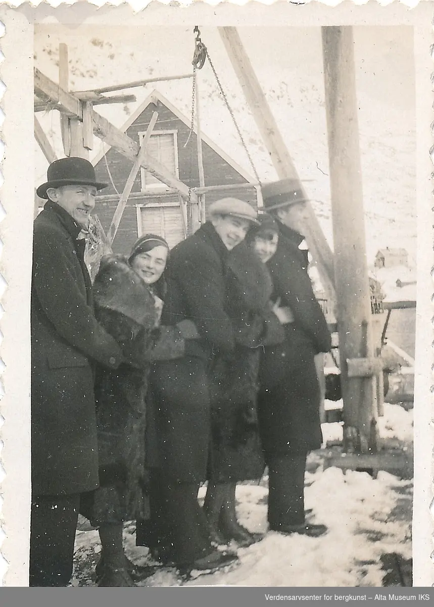 Fem mennesker, tre menn og to kvinner, står på en rekke og holder rundt hverandre på en kai. De ler og smiler, og de er kledd i vinterklær. Det er noen snøflekker på bakken rundt dem.