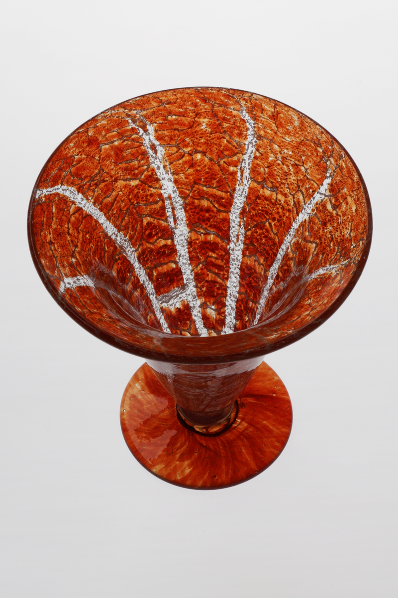 Konisk vase i Ikorakrystall som hviler på en sirkulær fot. Korpus har en marmoreringslignede overflate, hvor melkehvite og klare partier bryter opp den rødfargede bunnen.