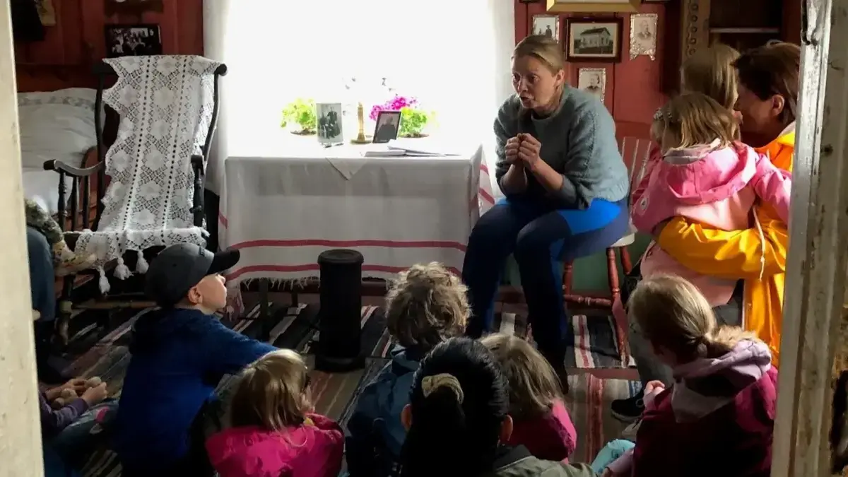 Lite rom i gammelt hus, med mye bilder og krimskrams på veggene. Barn sitter på gulvet og hører på dame som forteller historier.