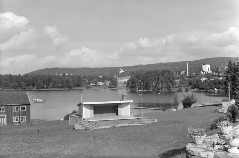 Svart-hvitt fotografi av utescene med litt overbygg og skråstilte tribuner foran, som ligger med ryggen mot Mjøsa. Løtenbygningen til venstre og danseplatting til høyre.