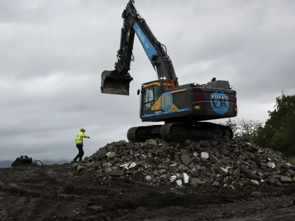 En blå og litt gul Volvo gravemaskin står på en haug av stein, i skarp silhuett mot grå himmel. En mann med gul refleksjakke og hjelm beveger seg opp mot maskina fra venstre.