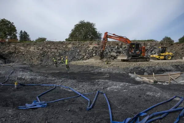 Tre arbeidere i signalklær og hjelm  jobber nede i "krateret" på byggetomta sammen med en oransje gravemaskin og en gul veivals. Mange meter med blå slange ligger i forkant i bildet.