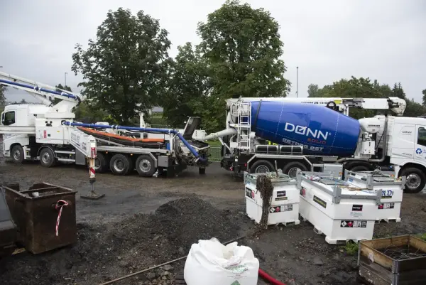 En hvit betongbil med blå betongtrommel forer en annen hvit bil med betong. Den andre hvite bilen har slangesystem som gjør det mulig å spre betongen over et stort område.