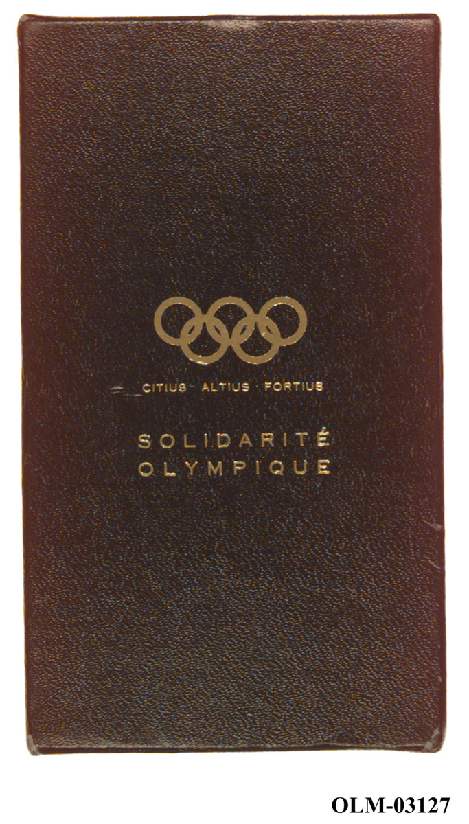 Gullfarget klype av metall med motiv av de olympiske ringene.
Det følger med en eske til klypen. 