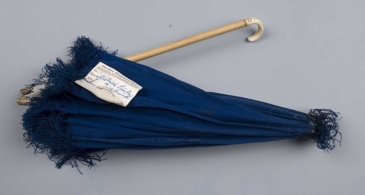 Liten blå parasoll med frynser langs kanten og på toppen. Foret med hvit silke. Spiler med pynteknotter. Stangen er todelt og låses med et messingbeslag, dekorert håndtak.