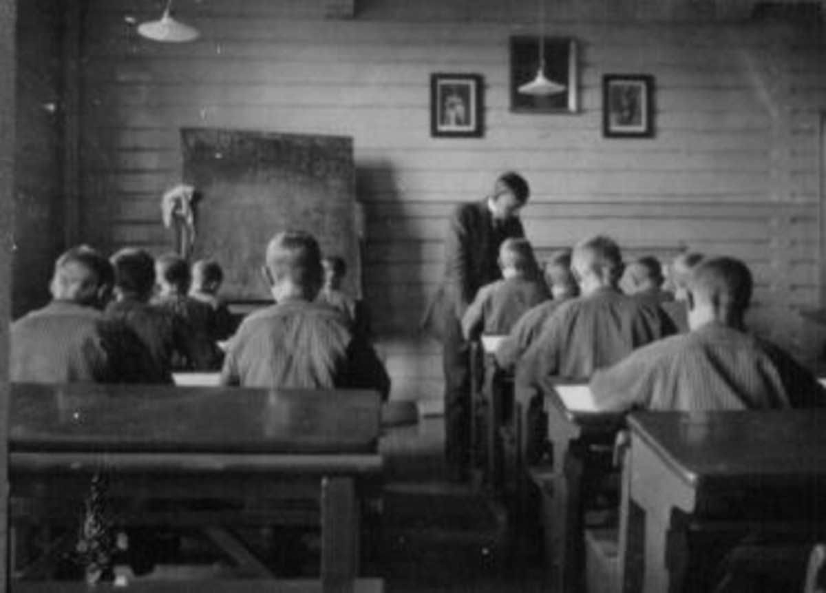 Interiør i klasserom på  Toftes Gave, Nedre Sund, Helgøya. Elever ved trepulter og lærer.