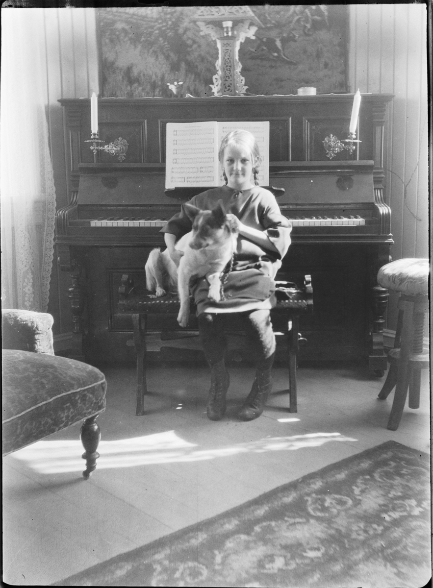 Interiør, stue. Jente med en hund på fanget sitter foran et piano. 