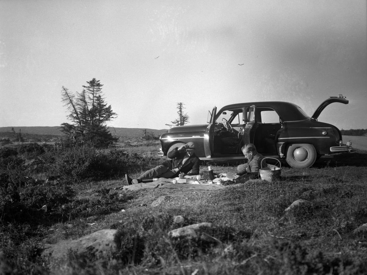 To menn tar ei kaffepause under en biltur på fjellet. Bilen er en Dodge eller Plymouth, ny modell i 1949 som gikk relativt uendret til 1952. 
