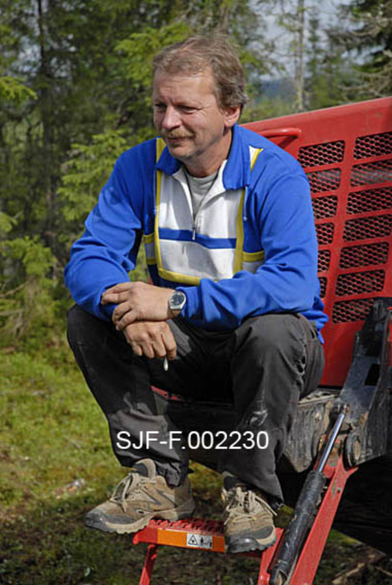 Maskinentreprenør Ola Engen fra Søre Osen i Trysil, fotografert sittende på trappa opp mot førerhuset på sin fire år gamle Valmet lassbærer.  Bak ham ser vi deler av motorkassa på maskinen.  Engen er iført joggesko, fritidsbukser og en blå bomullsgenser med kvite og gule striper.  I den ene handa holder han en sigarett.  Bildet er tatt i Ulvåsen i østre Elverum, 5. august 2009. 