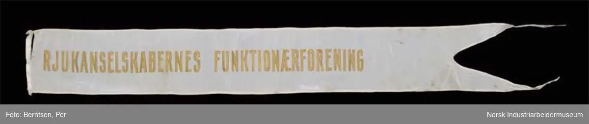 Silke banner for Rjukanselskapenes Funksjonærforening. Banneret er hvit og har gullfarget tekst på en side. Den splittes i to spisser i enden og har revne ved øvre feste.