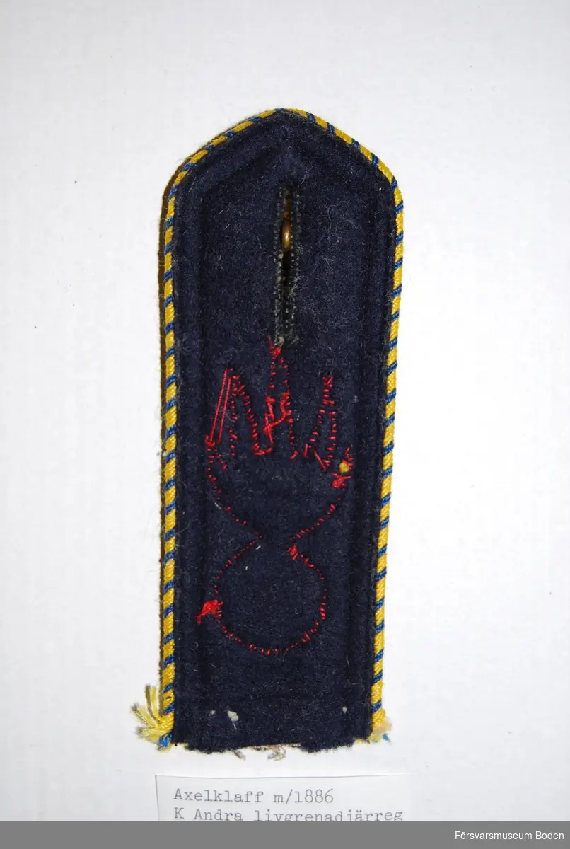 Mörkblått kläde med blått och gult dubbelt redgarnssnöre m/1873 runt kanten. Granat m/1873 av rött kläde för Andra livgrenadjärregementet (I 5) och slät knapp i guld upptill. Knappen är lös och är avsedd att vara fastsydd på vapenrocken.