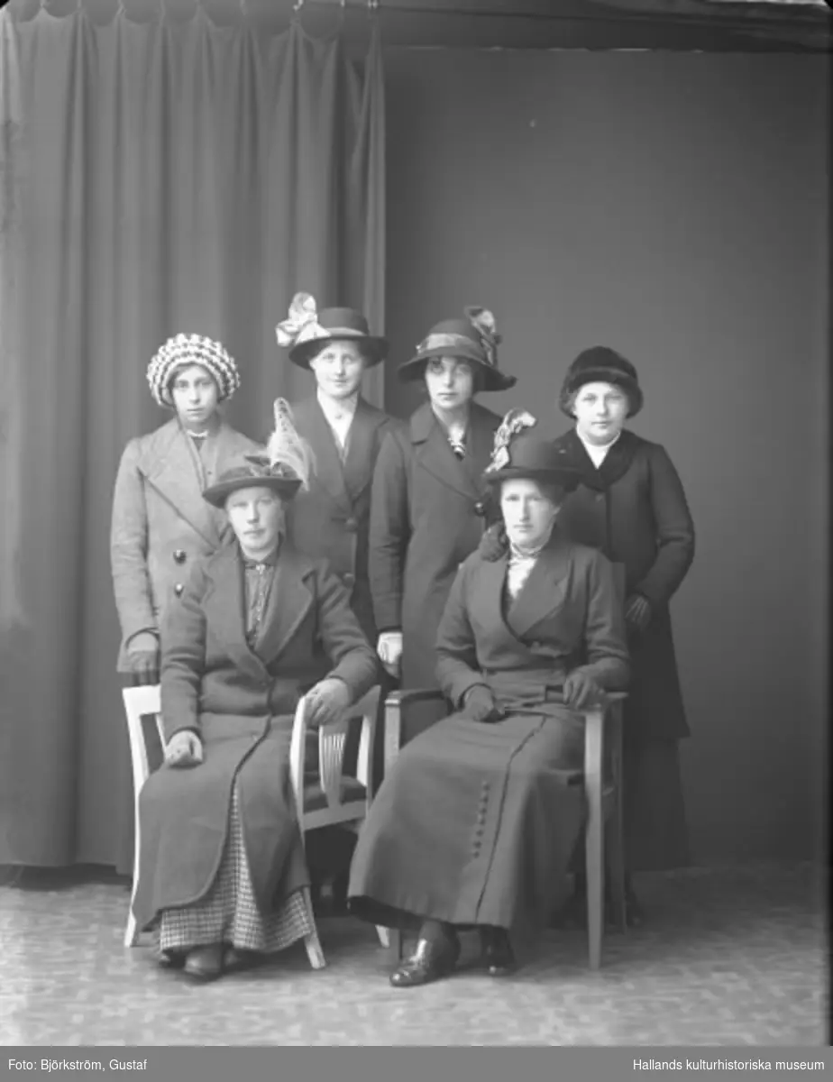 Ateljéfoto av sex kvinnor iklädda hattar och kappor. De sittande kvinnorna är systrar och heter Gerda Alfrida Johansdotter, som sitter till vänster, och Hilma (som gift sig i Tvååker till Andersson). Deras syster Hanna, gift Munther, står som nr 2 från vänster. Systrarna föddes i Veddige på Syllinge 7.