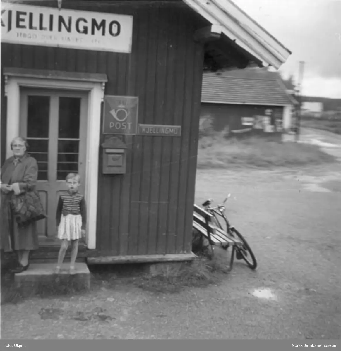 Kjellingmo stasjonsbygning, to mennesker utenfor stasjonsdøra, postskilt