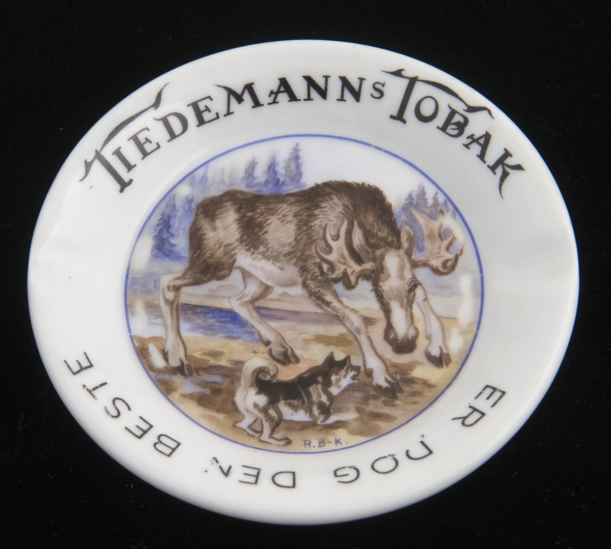 Lite, rundt askebeger i porselen med to fordypninger i randen. Påtrykket motiv av elg og elghund etter original av R. B-K.