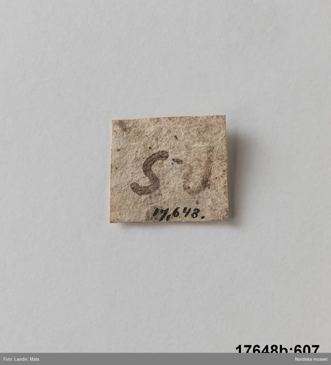 Pappersetikett (som ursprungligen har suttit på stenen). Fyrkantig med text på ena sidan "R S".